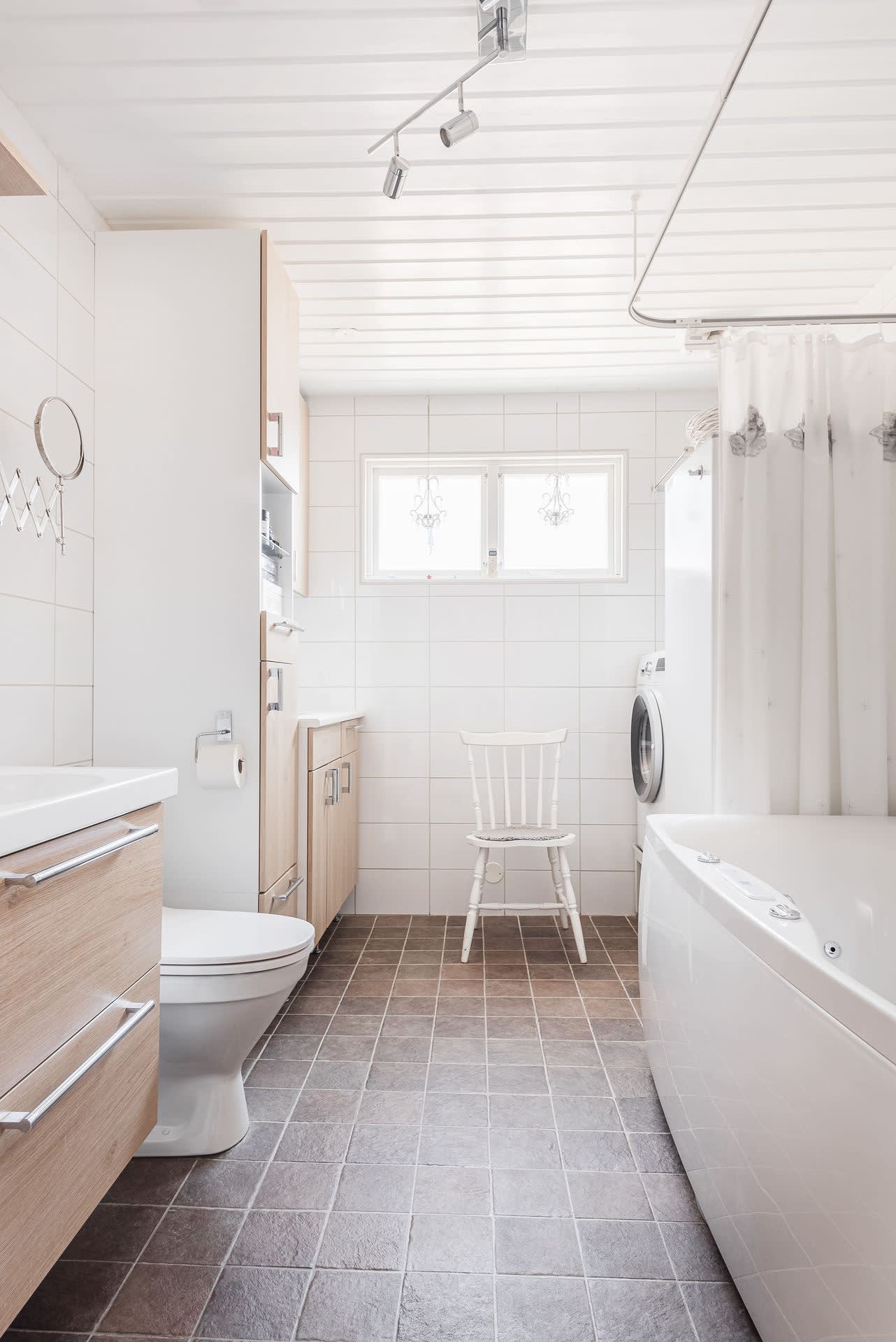 Helkaklat badrum/tvättstuga med bubbelbadkar, kommod och wc samt tvättmaskin, arbetsbänk med ho, varmvattenberedare, fönster och fläkt.